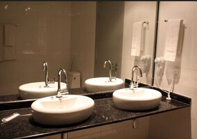 Un baño con tres lavamanos, cada uno con un espejo encima. Hay un perchero en la pared y un secador de pelo en la encimera.