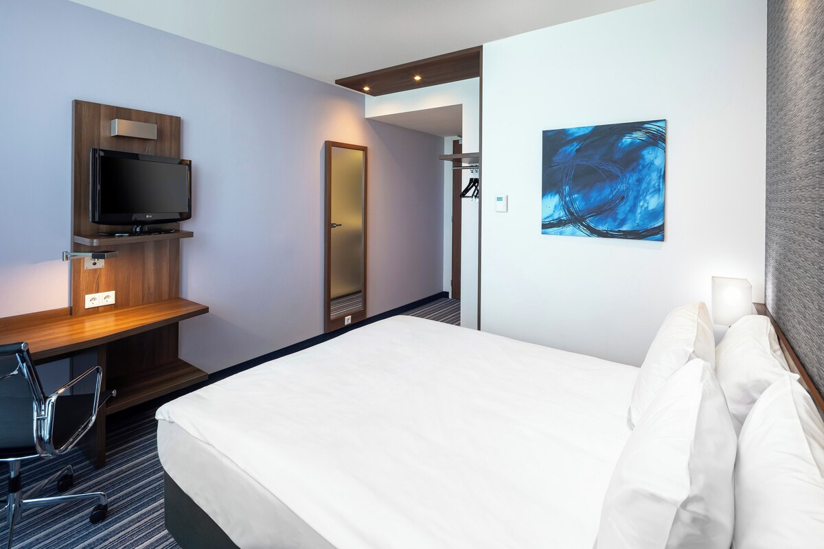 A hotel room with a bed, a desk with a flat-screen TV, a chair, a mirror, and a painting on the wall.