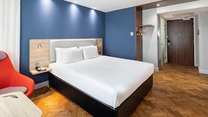 Una habitación de hotel moderna con una cama grande, una mesita de noche de madera, una silla roja y suelo de madera.