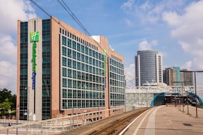 Un hotel moderno con un llamativo letrero verde, fachada de vidrio y un andén de estación de tren al fondo.