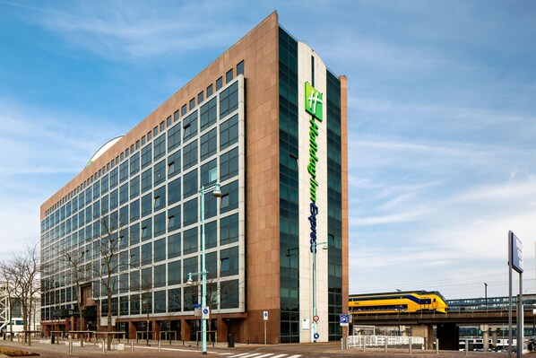 Un moderno edificio de hotel con un llamativo logo verde y blanco, amplias ventanas, y un tren pasando al fondo.