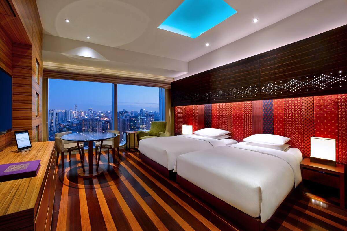 Quarto de hotel com vista para a cidade, uma escrivaninha com computador, uma mesa redonda, duas camas e uma parede com padrão vermelho.