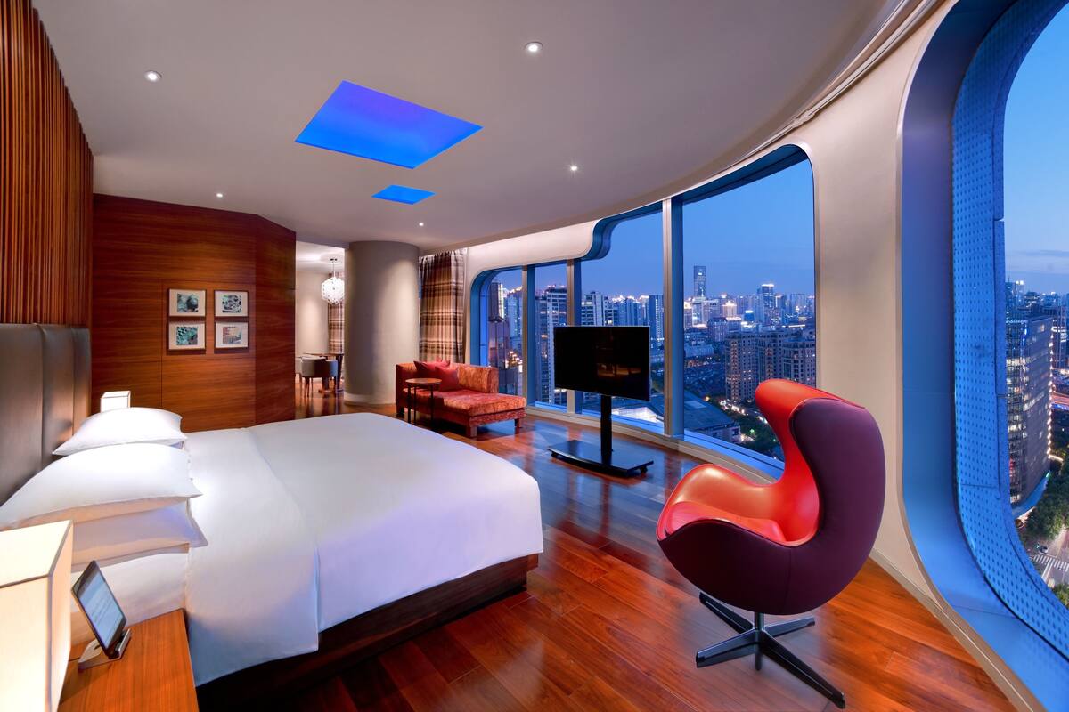 Quarto de hotel moderno com uma cama grande, uma TV, uma cadeira vermelha e vista para a cidade.