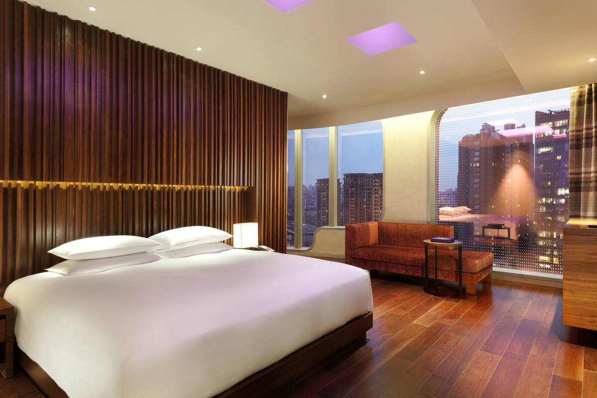 Quarto de hotel moderno com uma cama grande, área de estar com sofá e poltrona, e vista para a cidade através de grandes janelas.