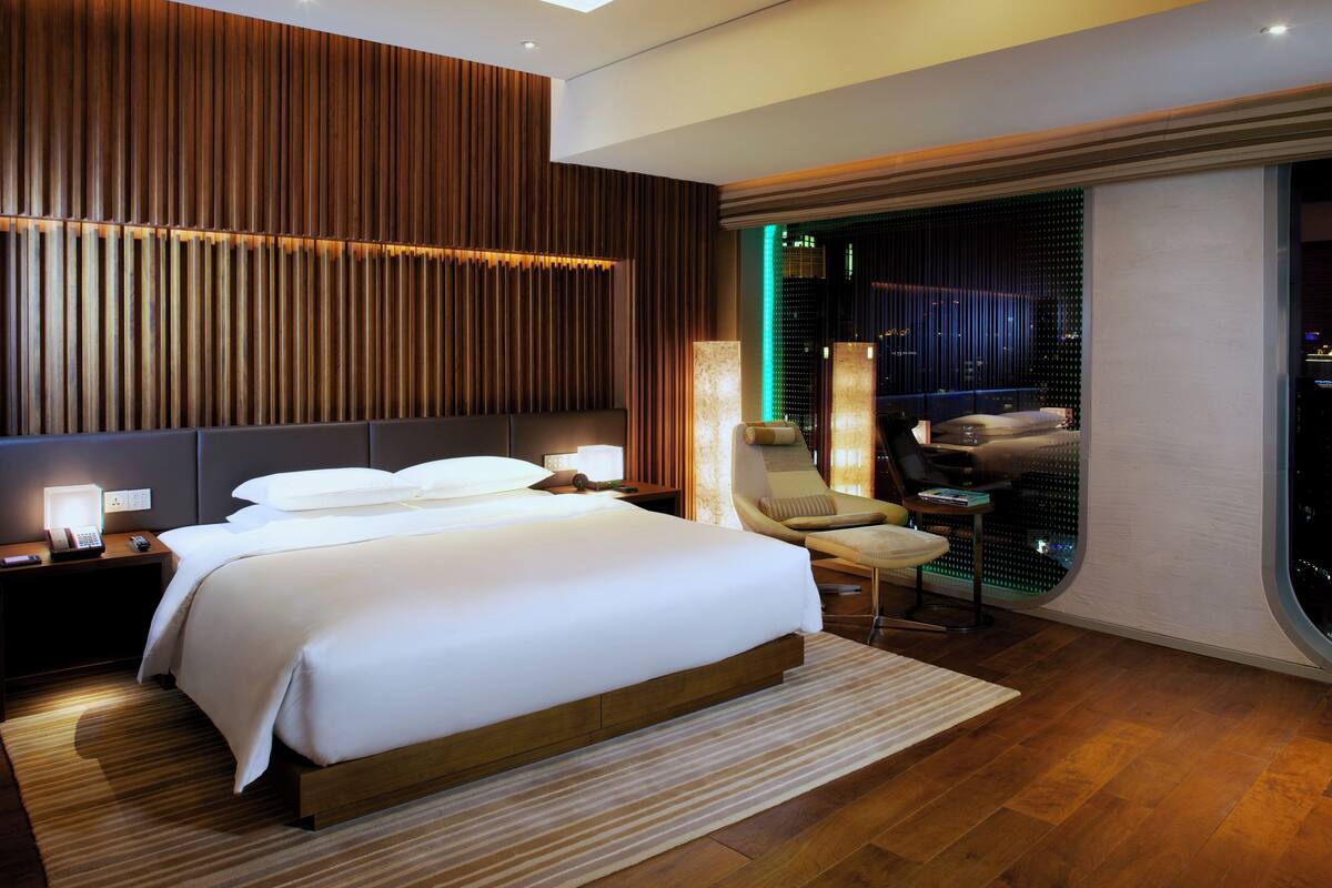Quarto de hotel moderno com uma cama grande, um espaço de estar com uma poltrona e uma mesa, e vista através de uma janela ampla.