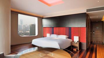 A modern hotel room with a large bed, a city view, a round dining table, and a unique ceiling design.