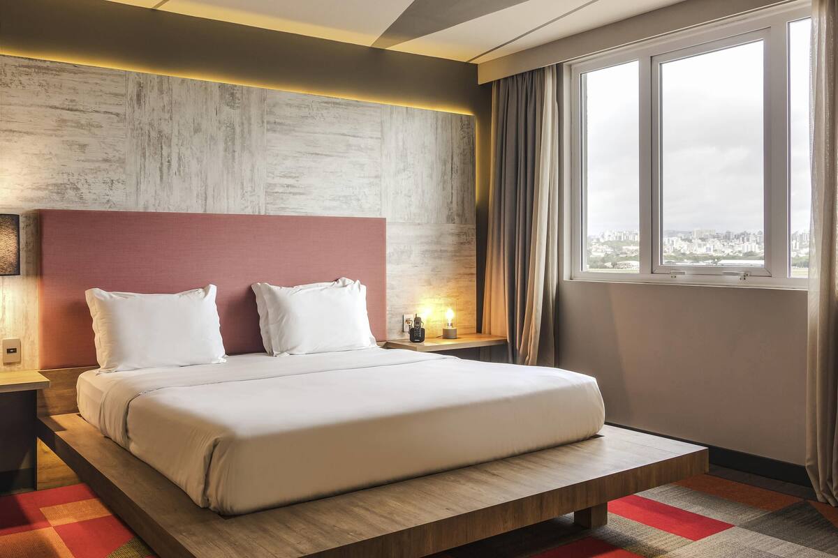 Habitación de hotel con una cama grande, cabecera de madera, mesita de noche, lámpara de noche y ventana con vistas a la ciudad.