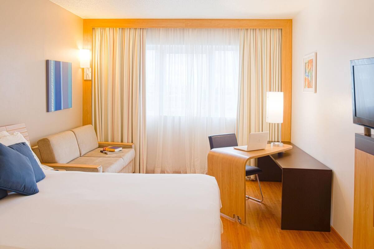Habitación de hotel con cama, escritorio con computadora portátil, silla, sofá, televisor y cortinas.