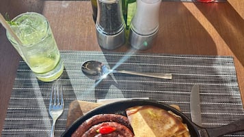 Un plato con salchichas y pan plano en una sartén negra, un vaso de bebida con pajita, saleros y pimenteros, y un tenedor sobre un mantel individual.
