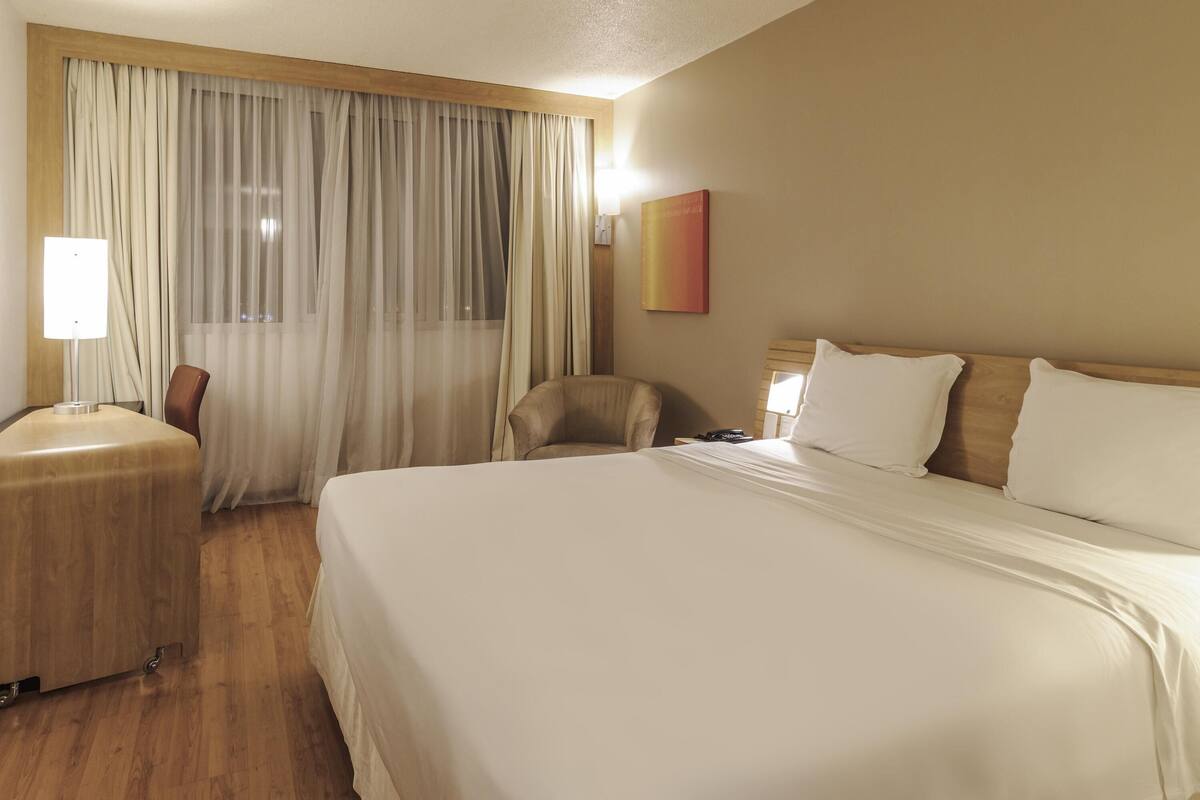 Habitación de hotel con una cama grande, una silla, un escritorio con lámpara y una ventana con cortinas.