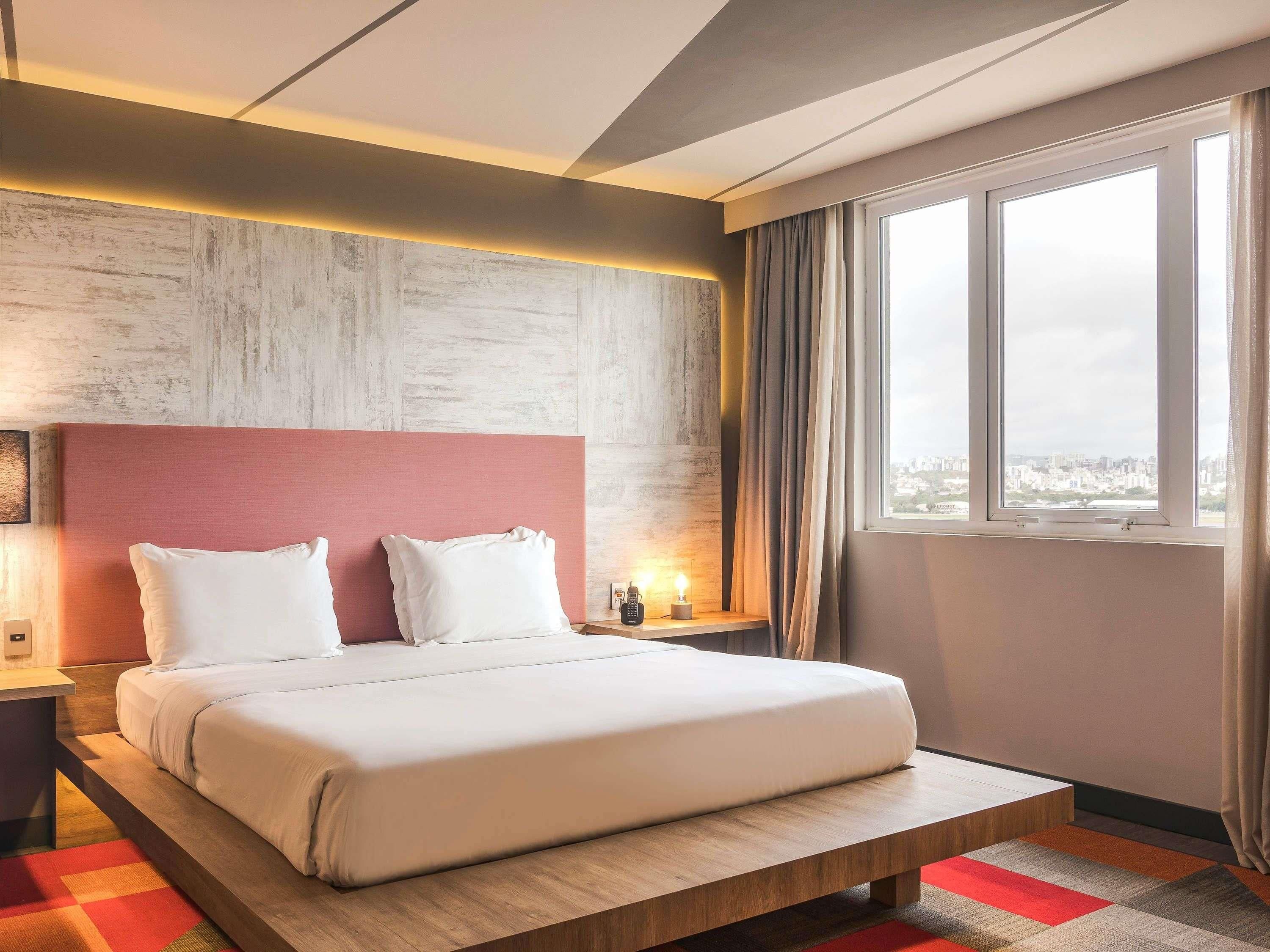 A hotel room with a bed, a nightstand, a window with curtains, and a city view.