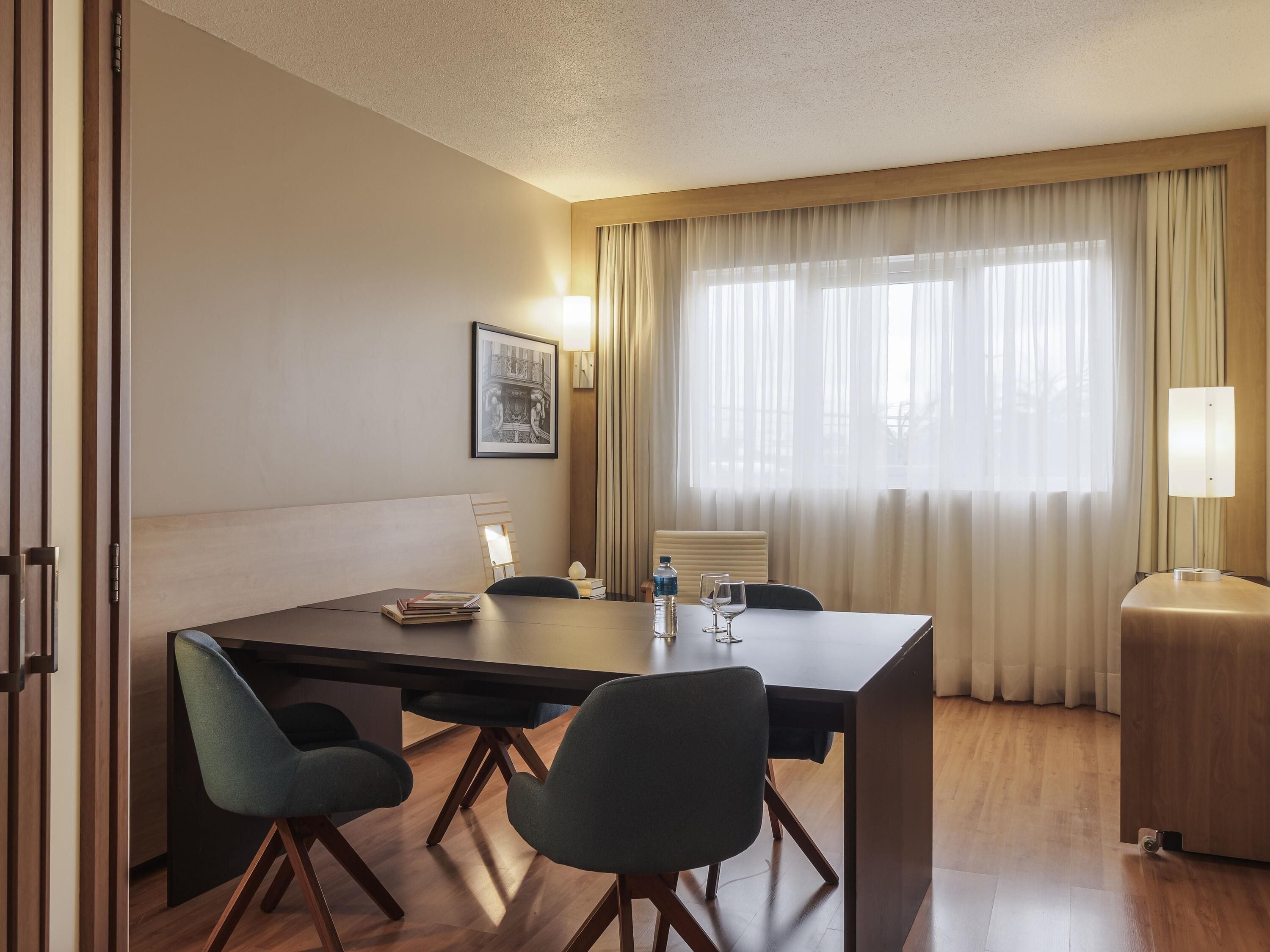 A hotel room with a wooden table, two chairs, a bed, a window with curtains, a wall-mounted lamp, and a framed picture.