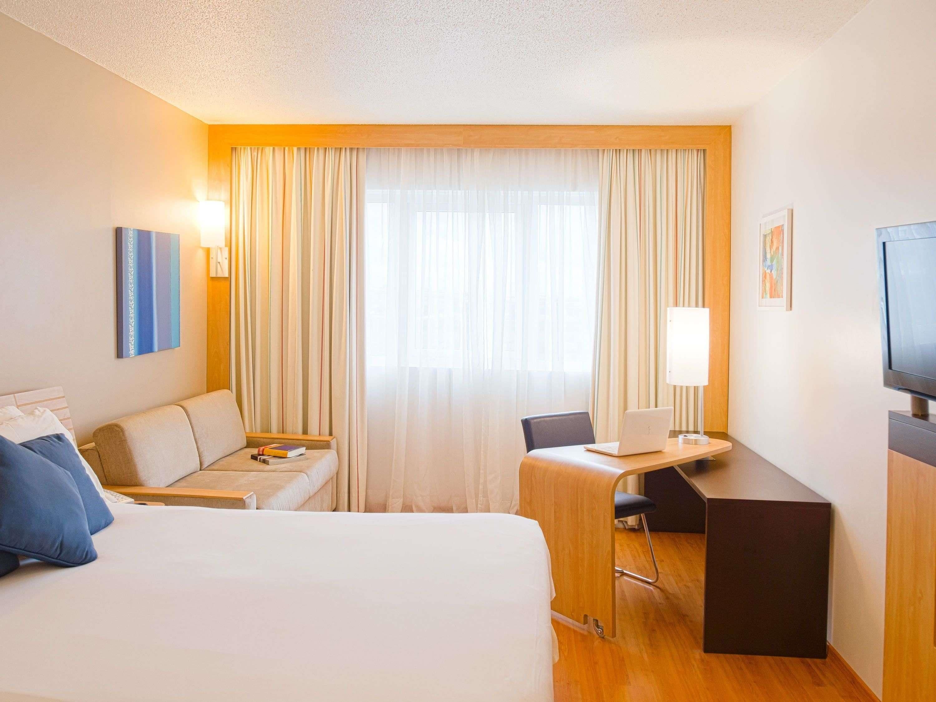 Habitación de hotel con cama, escritorio con una computadora portátil, una silla, sofá, televisor y cortinas.