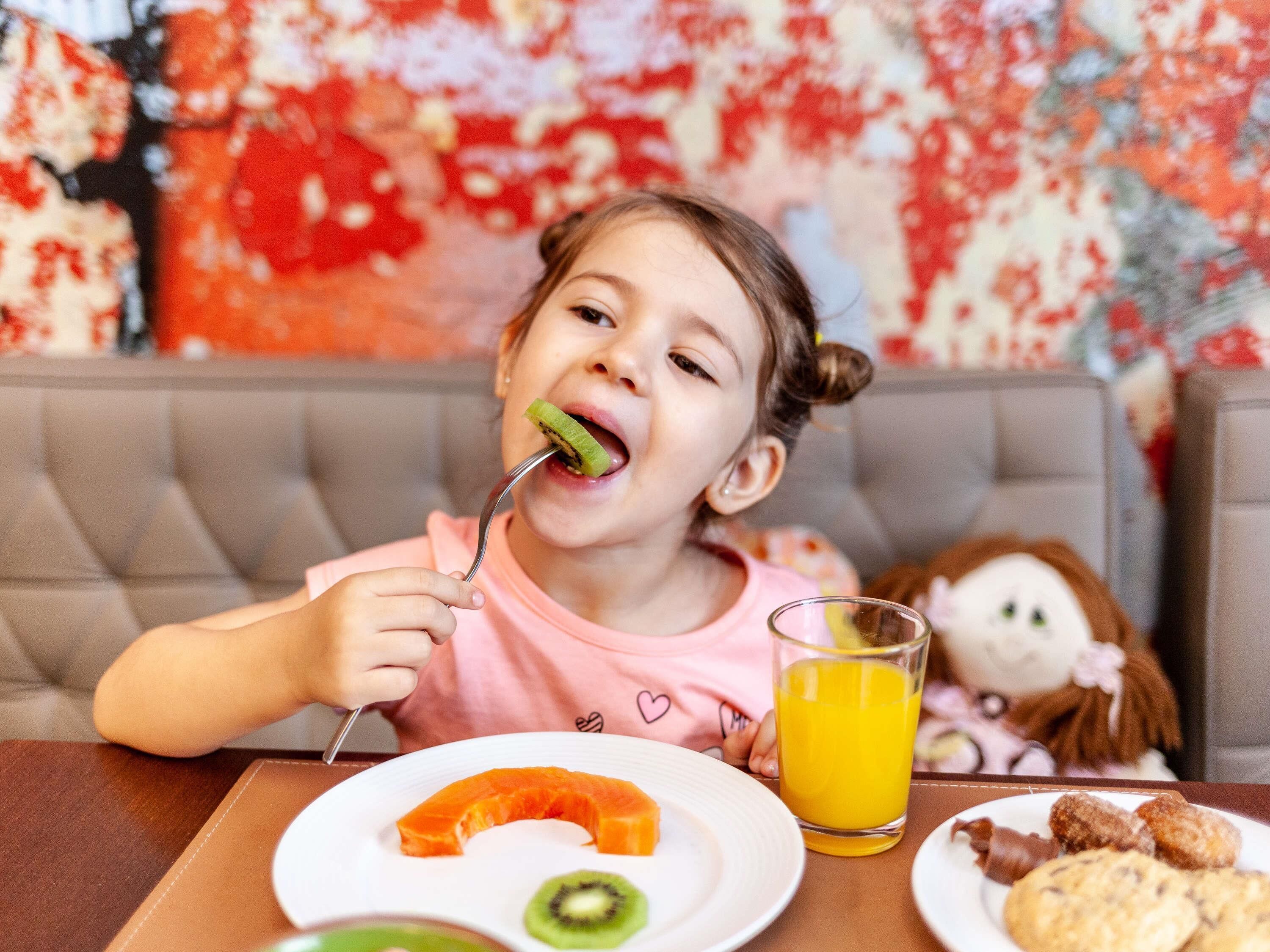A child sitting at a table with a plate of food, a glass of orange juice, and a stuffed toy in the background.