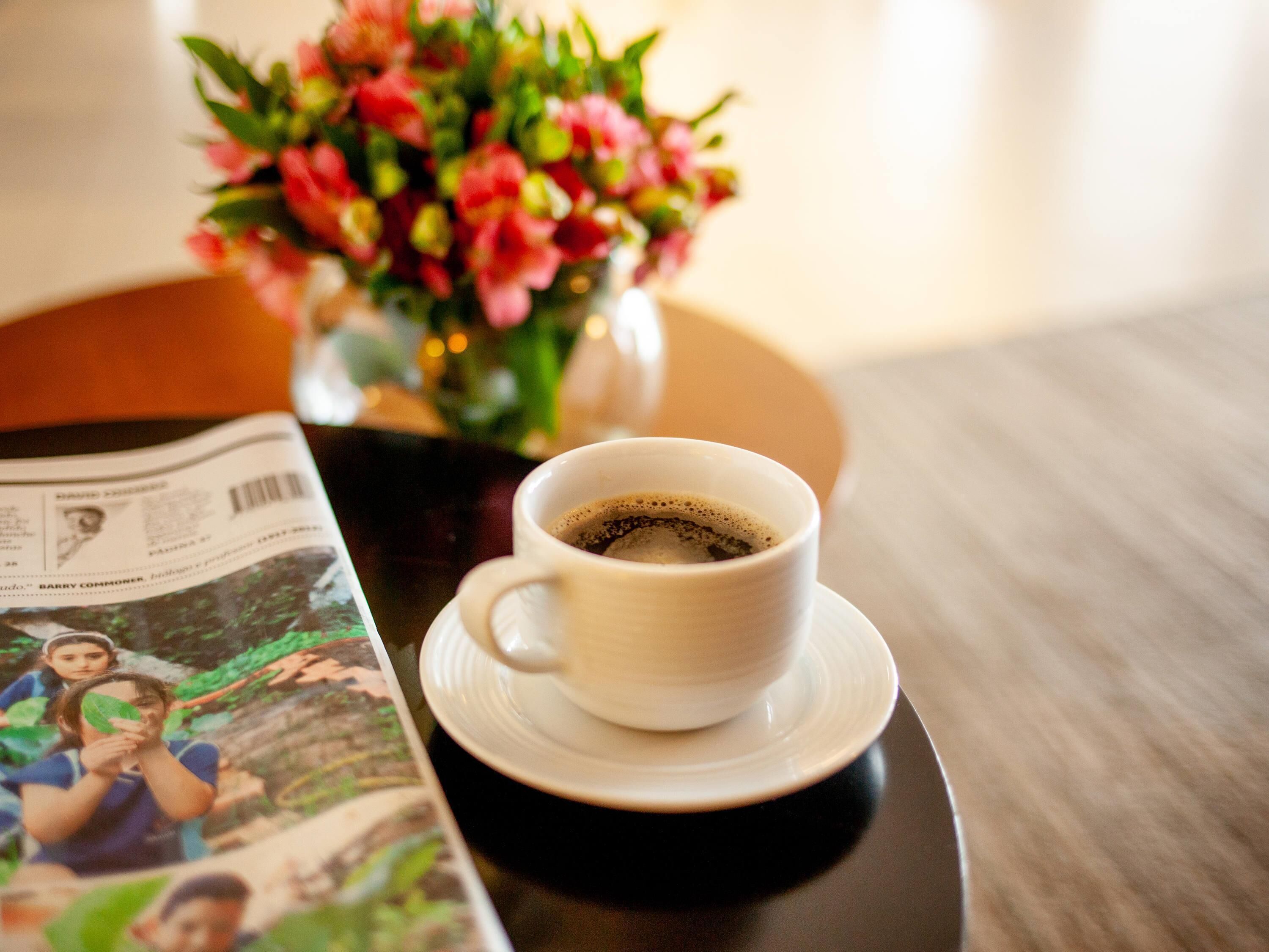 Una taza de café sobre un plato, una revista y un jarrón con flores sobre una mesa.