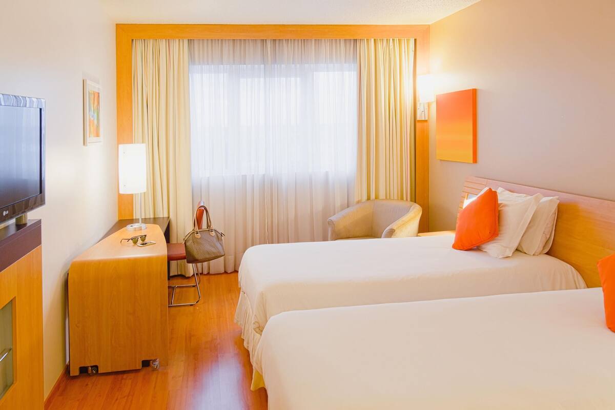 Habitación de hotel con dos camas, un escritorio, una silla, un televisor y una ventana con cortinas.