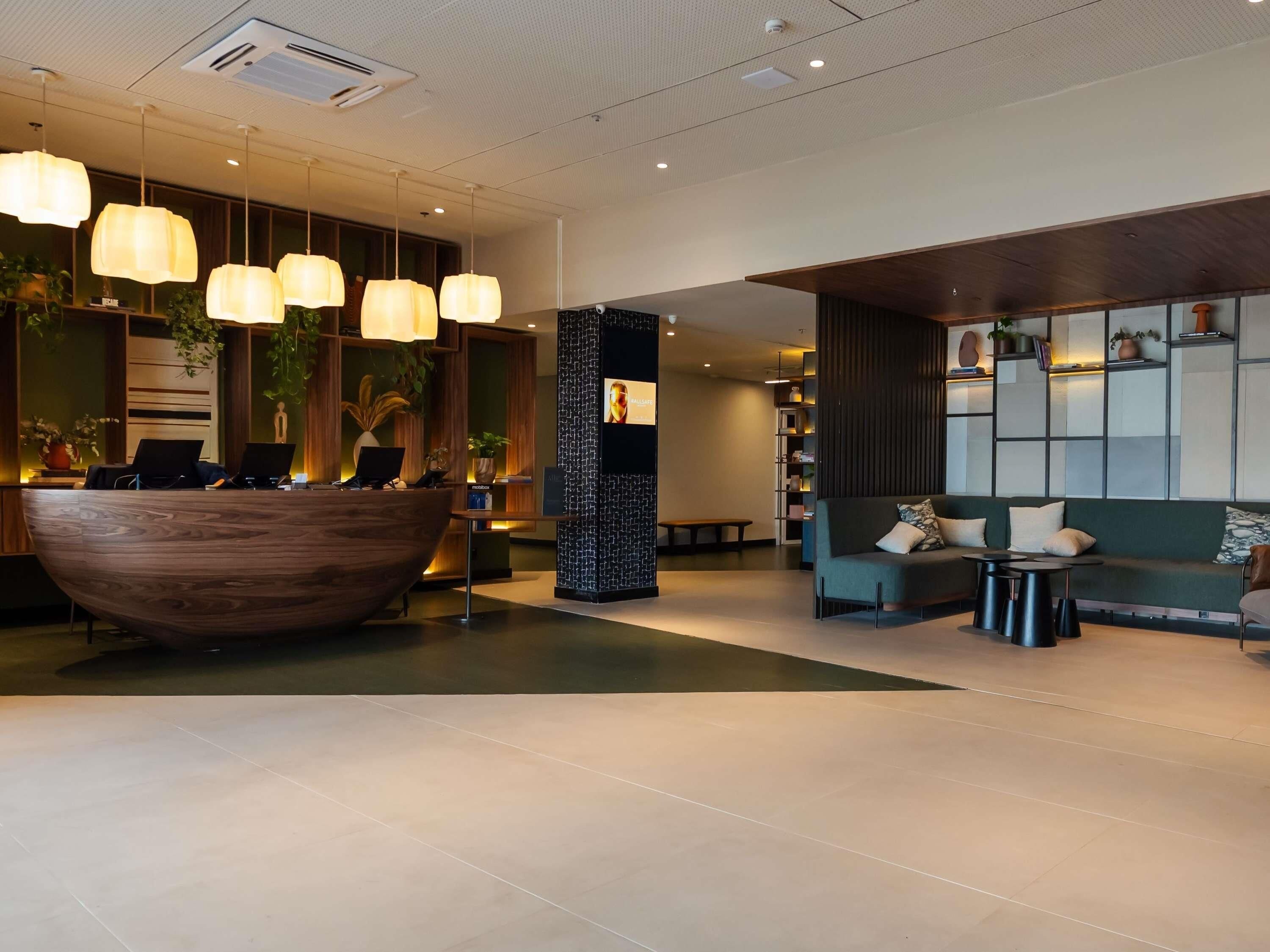Un área de recepción moderna con un mostrador de madera, lámparas colgantes y una zona de estar con un sofá y pequeñas mesas.