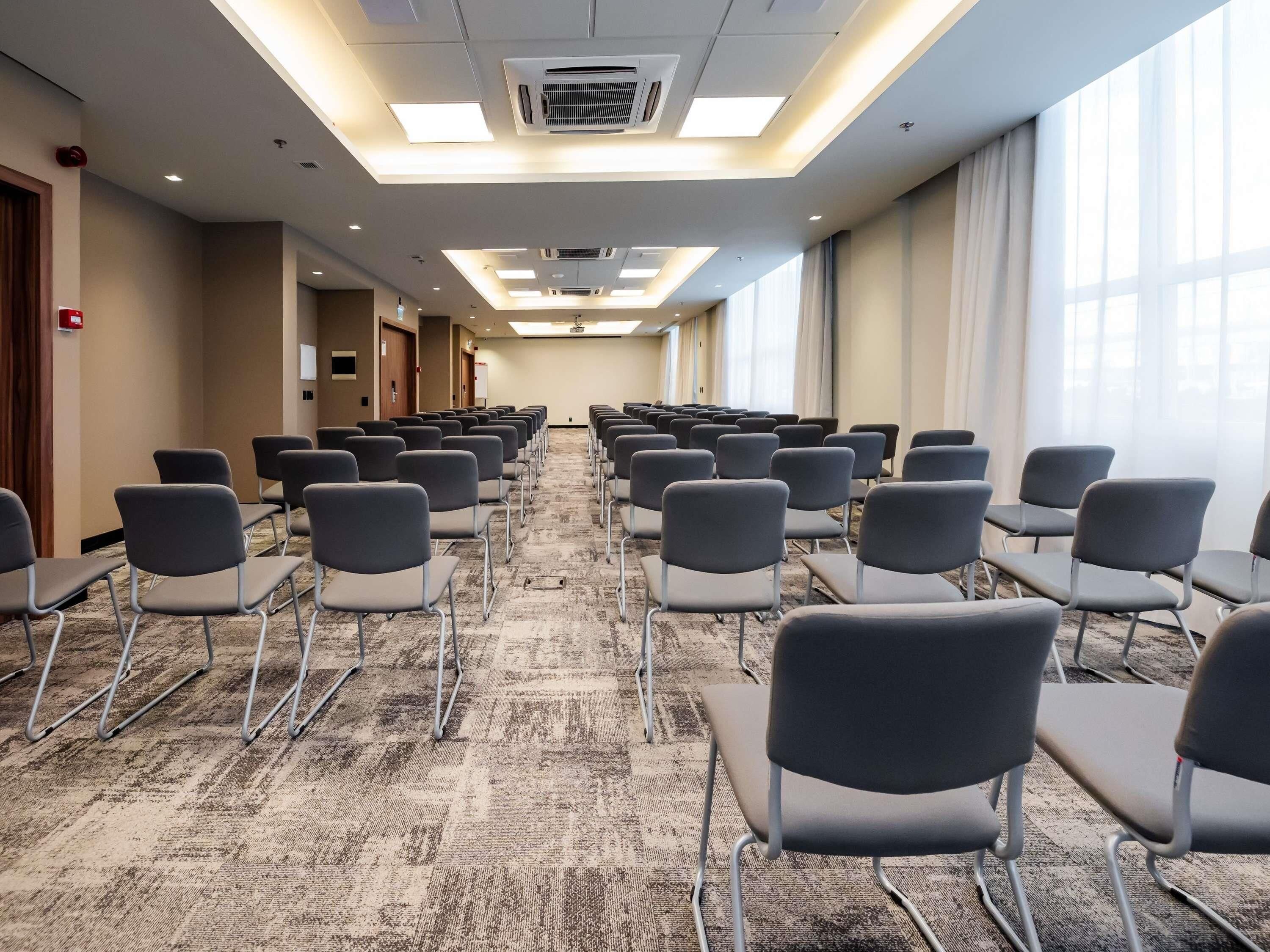 Un salón de conferencias con filas de sillas, piso alfombrado y ventanas amplias.