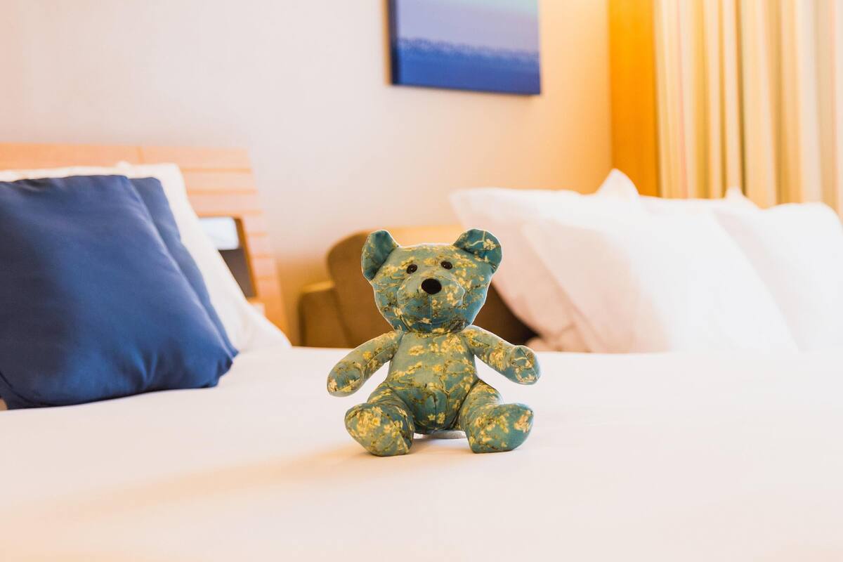Una cama con ropa blanca, una almohada azul y un pequeño osito de peluche azul sobre la cama.