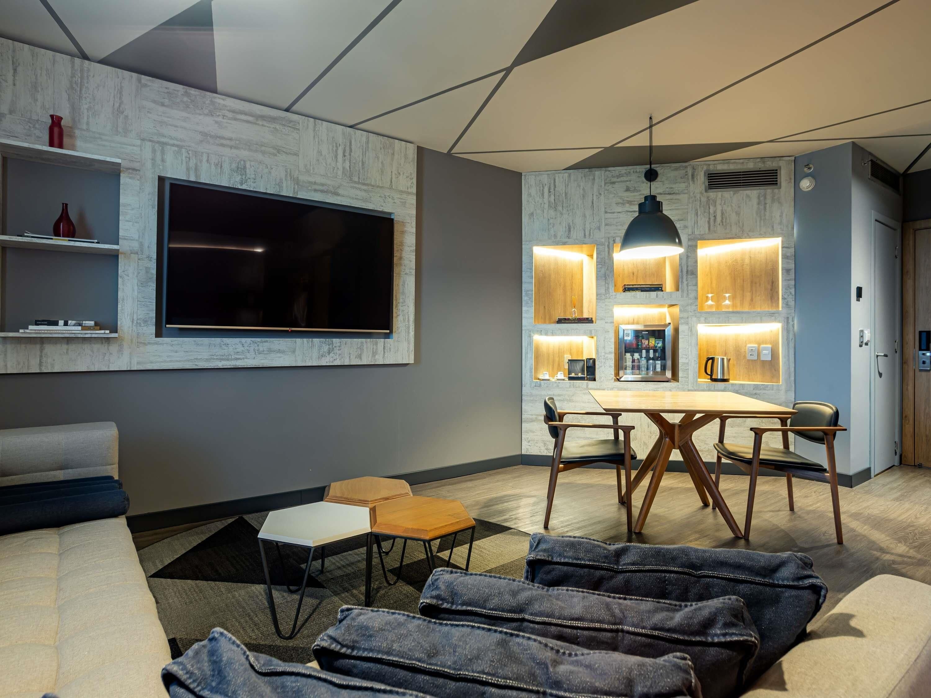 A modern living room with a flat-screen TV, a dining area with a wooden table and chairs, and a kitchen area with built-in lighting.