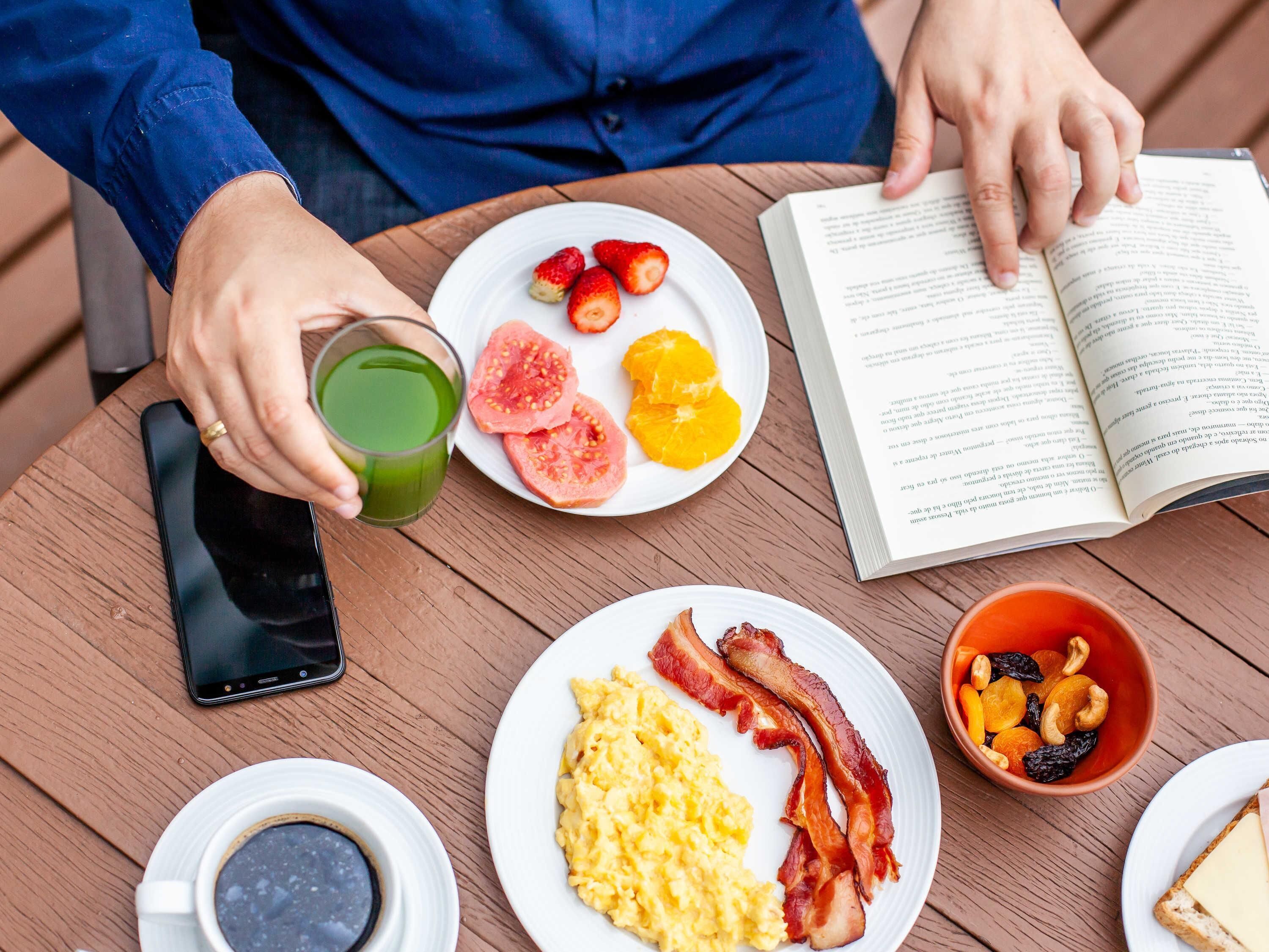 Un desayuno con un libro, un vaso de jugo verde, huevos revueltos, tocino y un tazón de nueces.