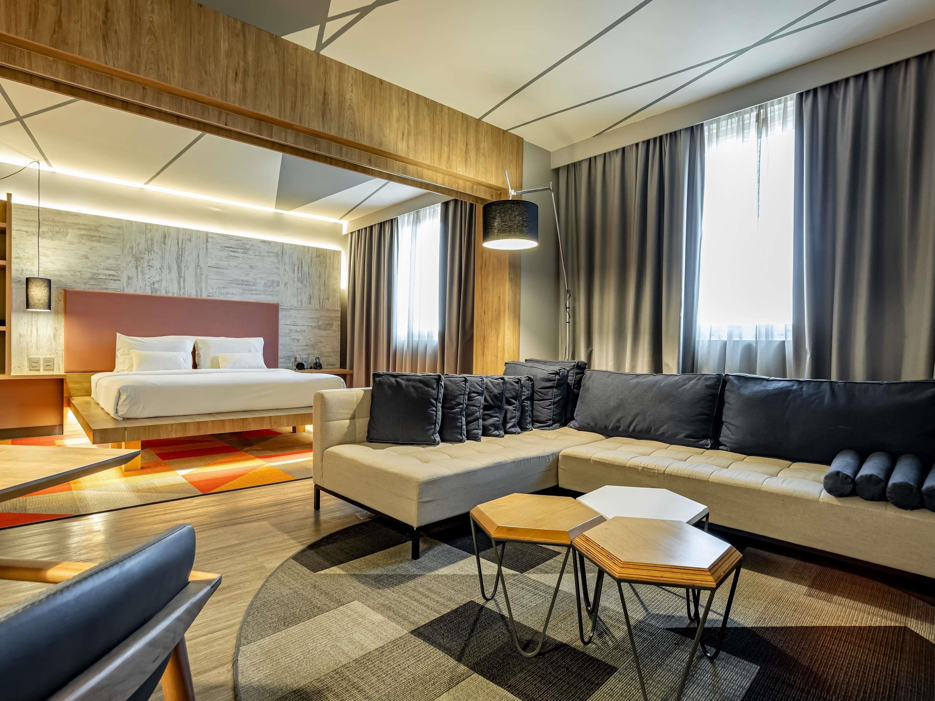 Een moderne hotelkamer met een bed, een slaapbank, twee nachtkastjes en een groot raam met gordijnen.