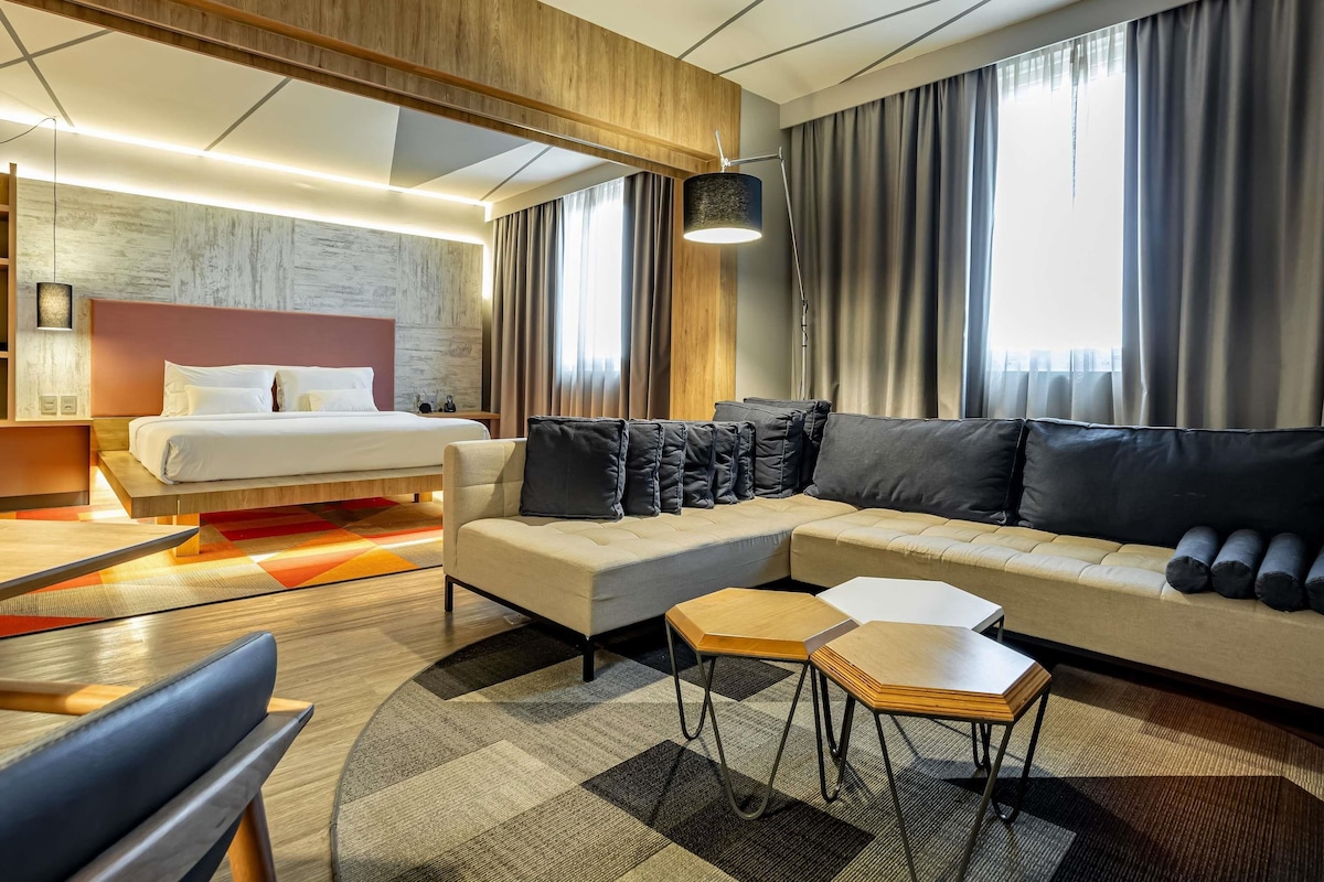 A modern hotel room with a bed, sofa, two side tables, and a large window with curtains.