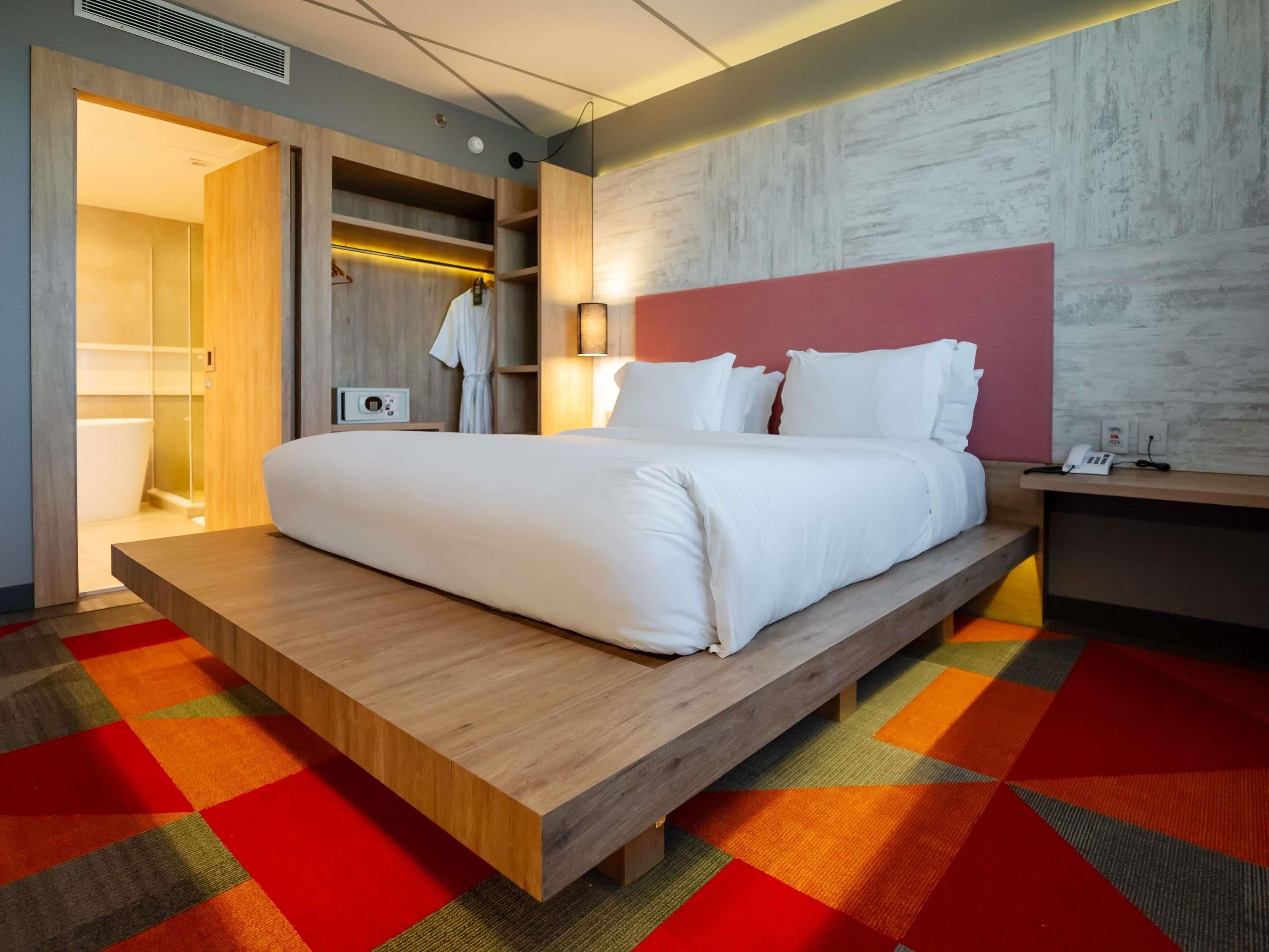 A modern hotel room with a large bed, a wooden platform, a colorful carpet, a wardrobe, and a bedside table with a lamp.