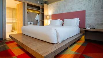 A modern hotel room with a large bed, a wooden platform, a colorful carpet, a wardrobe, and a bedside table with a lamp.
