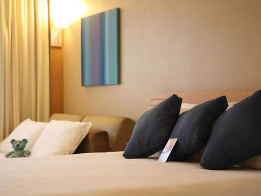 A hotel room with a bed, pillows, a teddy bear, and a painting on the wall.
