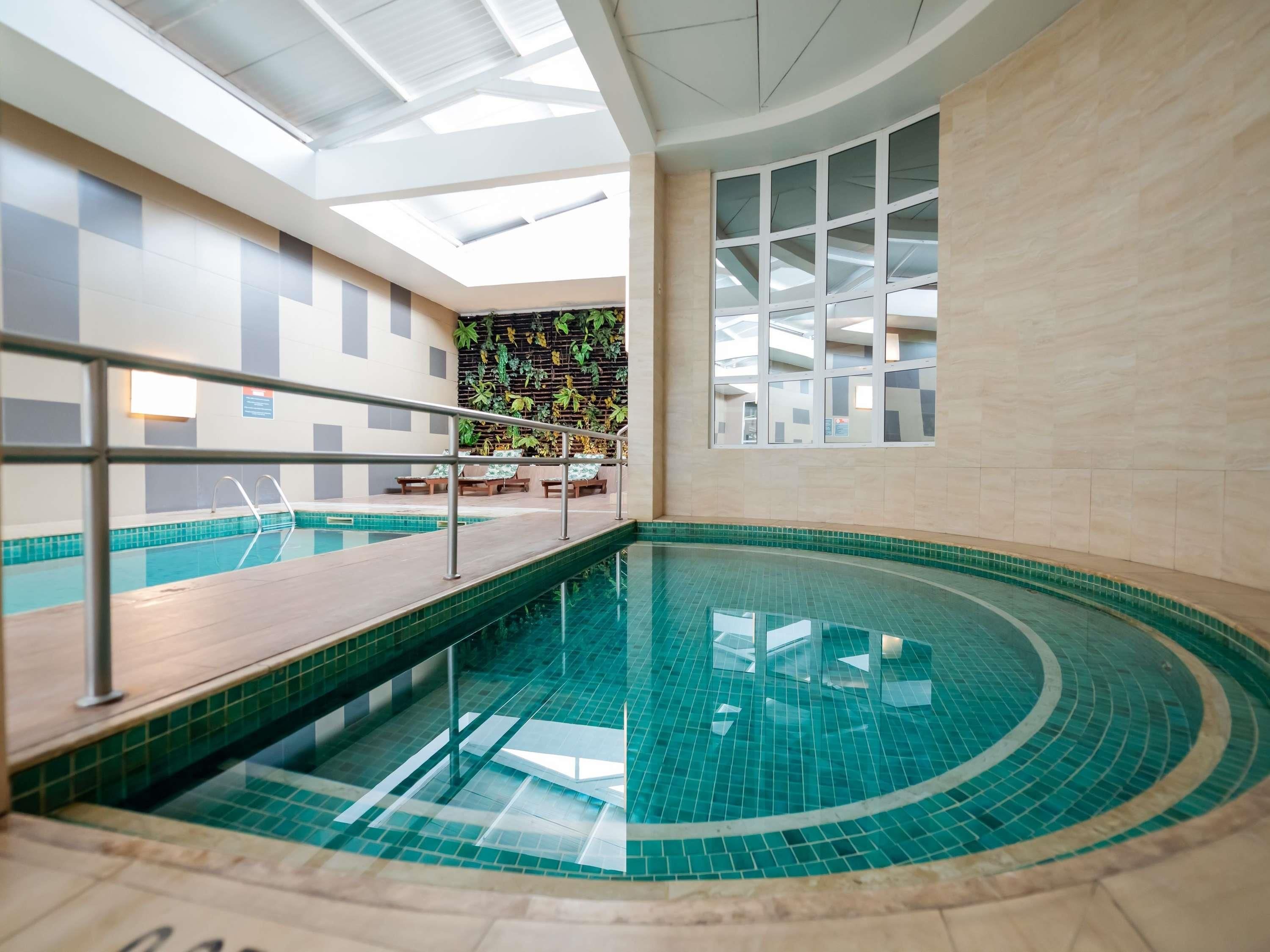 Una piscina interior con piso de vidrio, cubierta de madera y zona de estar con sillas y una mesa.