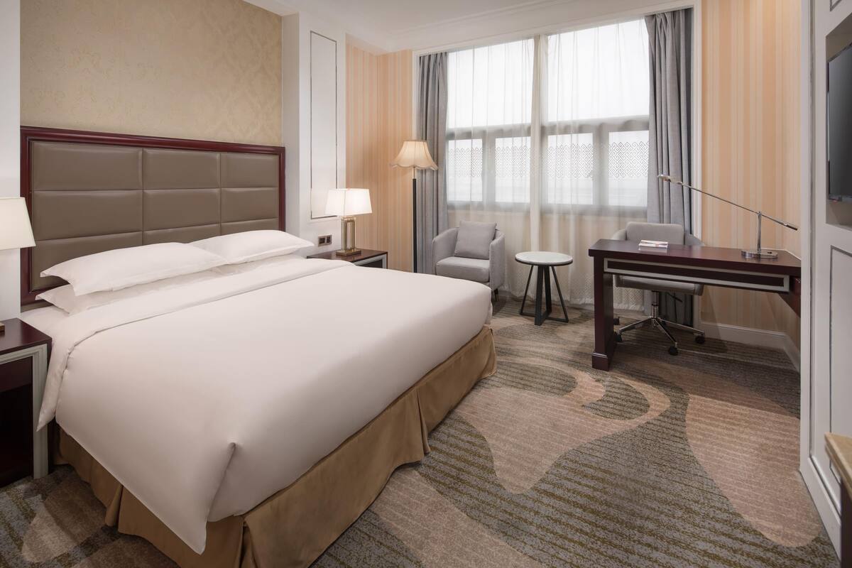 Et hotelværelse med en stor seng, et skrivebord, en stol, et lille bord og et fjernsyn.