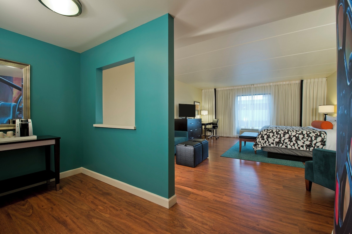 A hotel room with teal walls, a wooden floor, a bed with a patterned bedspread, a desk with a computer, a television, and a coffee machine.