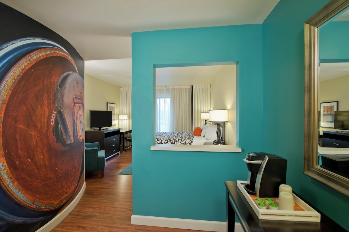 A hotel room with a teal wall, a bed with a patterned bedspread, a desk with a computer, a chair, a coffee machine, and a large painting on the wall.