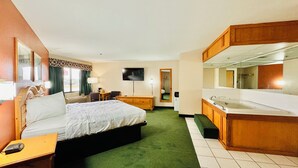 A hotel room with a bed, desk, chair, TV, and a bathroom with a sink and mirror.