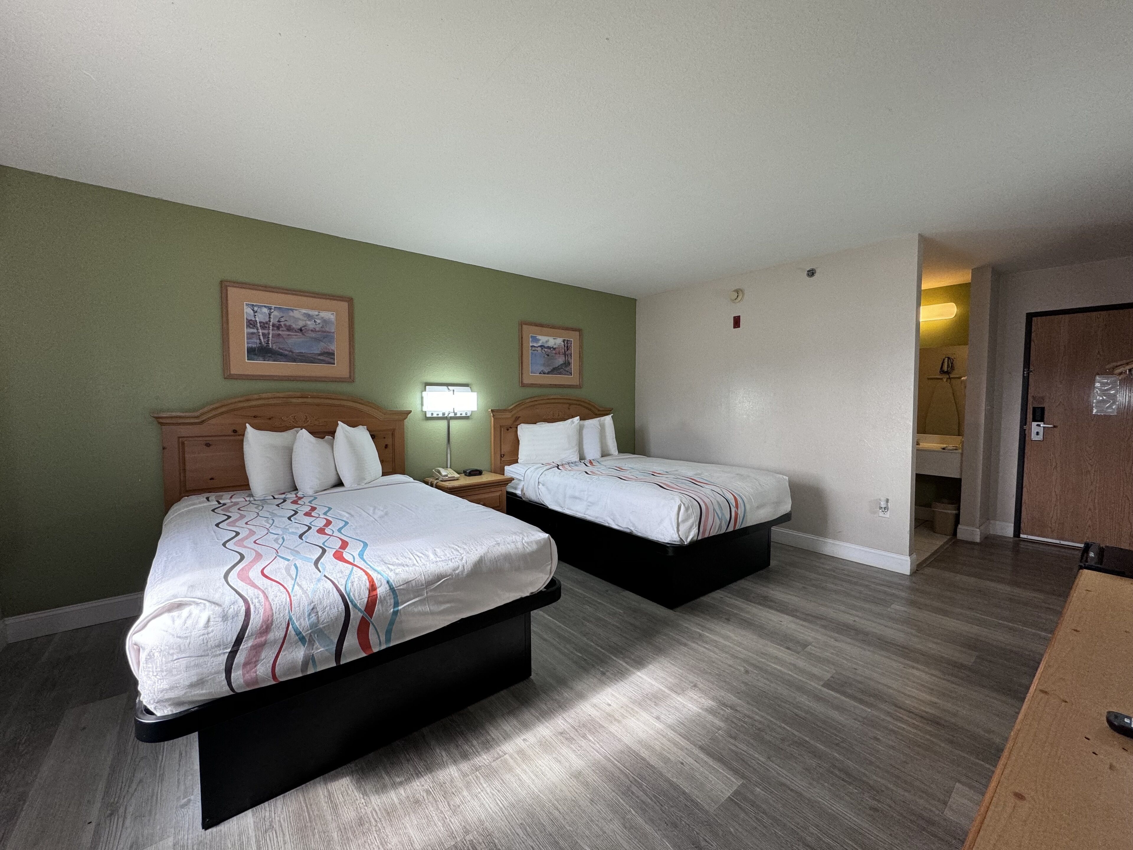 Una habitación de hotel con dos camas, un cabecero de madera, una mesita de noche con lámpara y un baño visible a través de una puerta abierta.