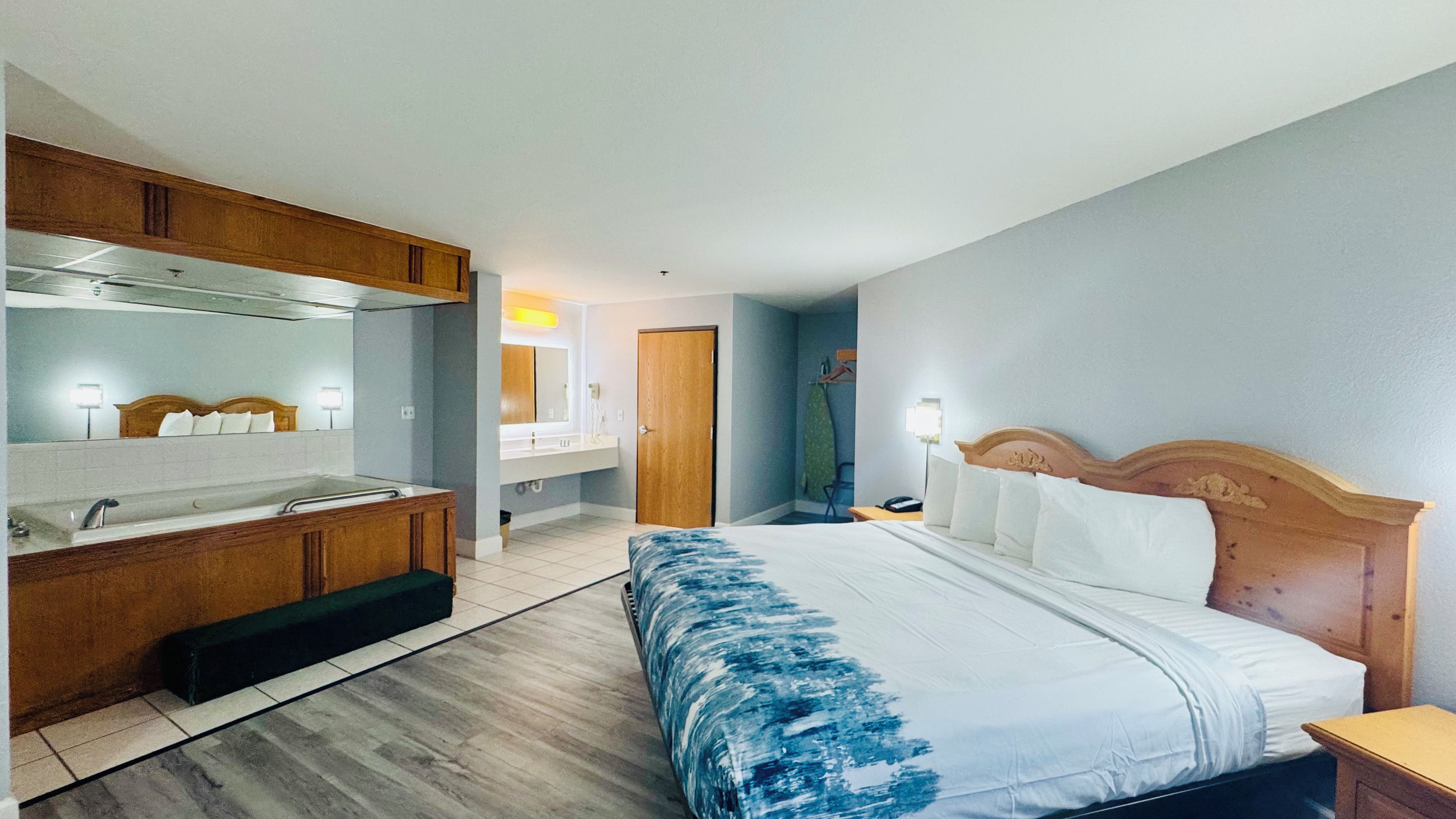 Une chambre d’hôtel comprenant un lit, une tête de lit en bois, une table de chevet, une commode, une salle de bain avec un lavabo et un miroir, et un banc en bois.