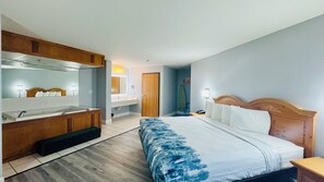 A hotel room with a bed, a wooden headboard, a nightstand, a bedside table, a bathroom with a sink and mirror, and a wooden bench. - Regency Inn And Suites (Stilwell)