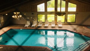 An indoor swimming pool with clear water, surrounded by white lounge chairs and large windows offering views of greenery.