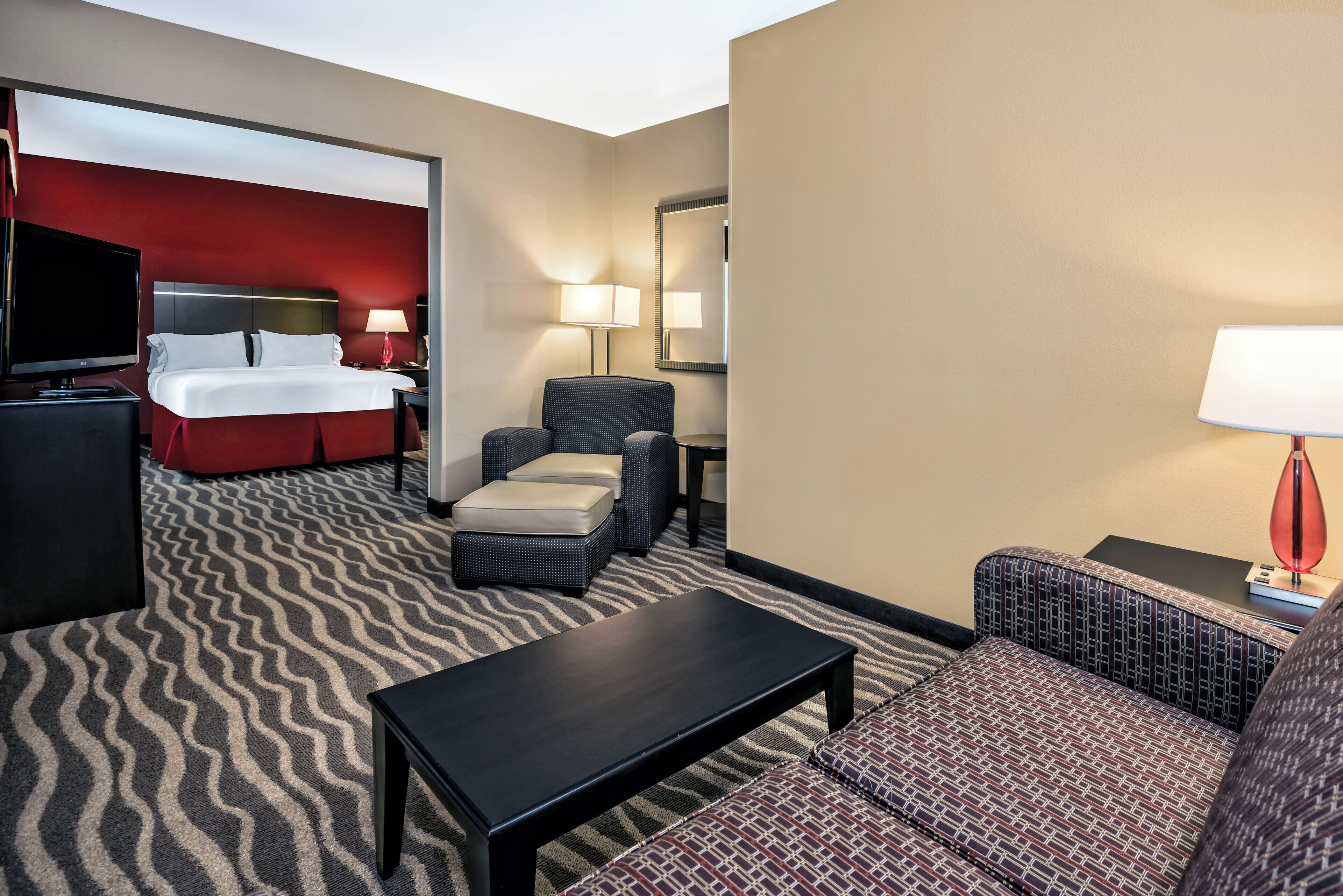 A hotel room with a bed, a TV, a desk, a chair, a sofa, and a patterned carpet.