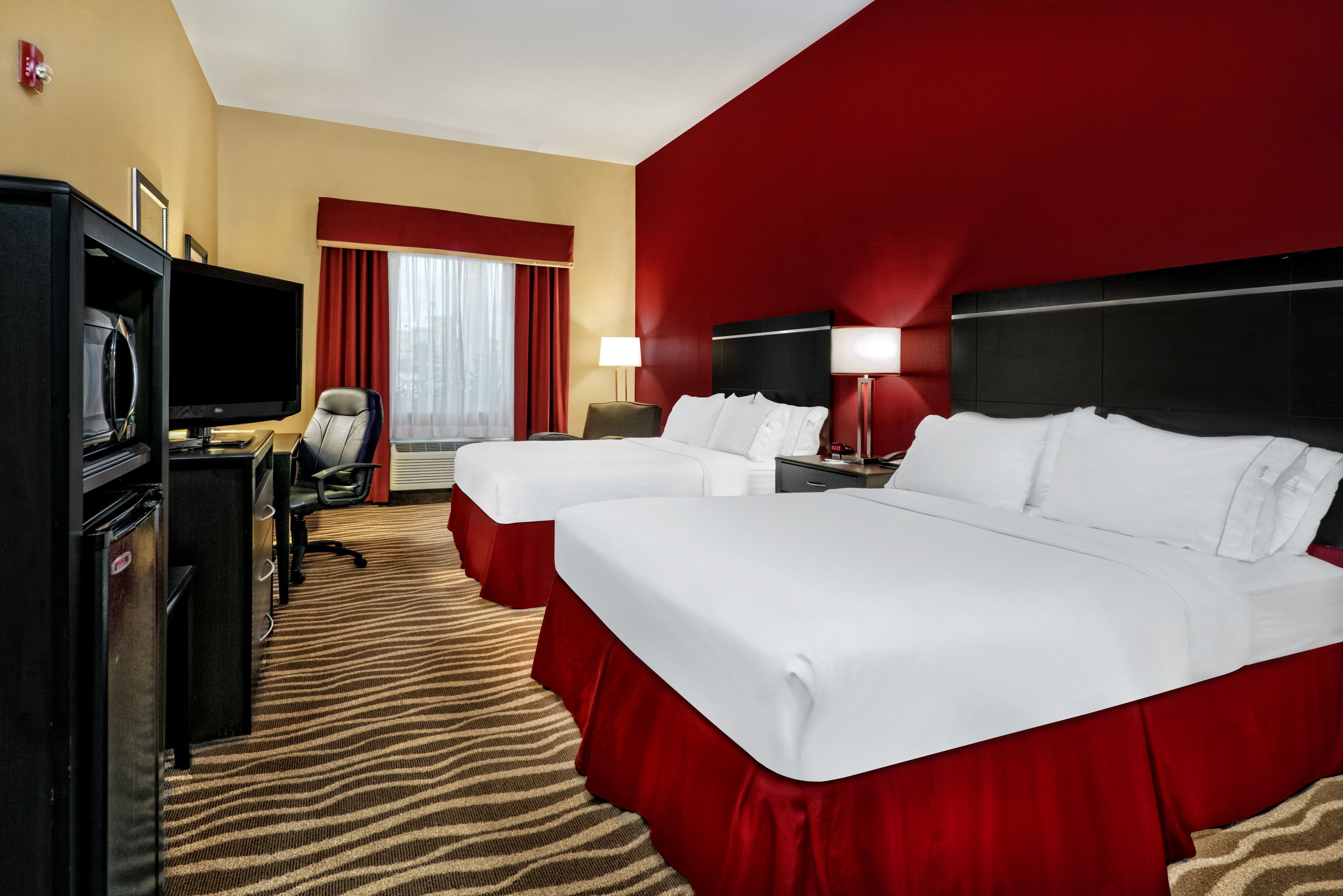 A hotel room with two beds, a desk with a television, a chair, and a red carpeted floor.