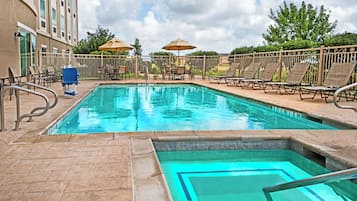 A swimming pool with lounge chairs and umbrellas, surrounded by a fence and a building.