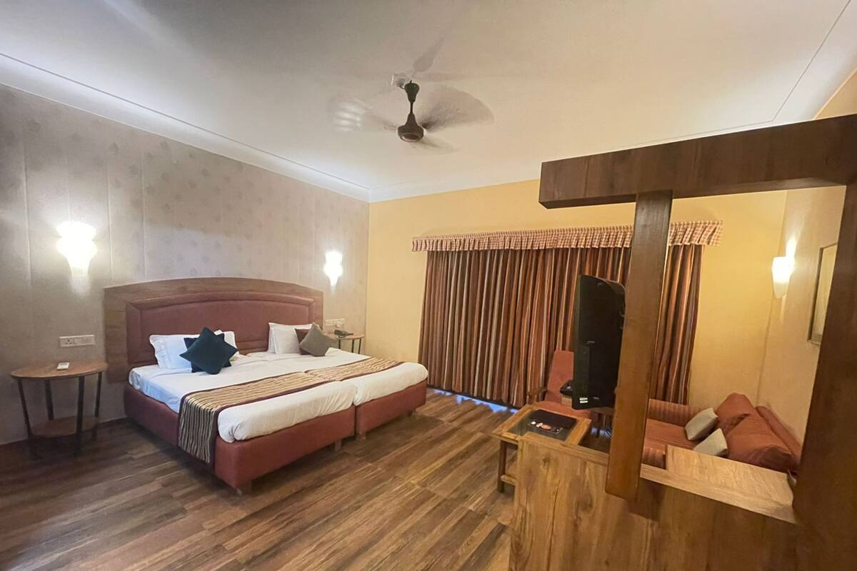A hotel room with a large bed, a wooden side table, a ceiling fan, and a view of the outdoors through curtains.