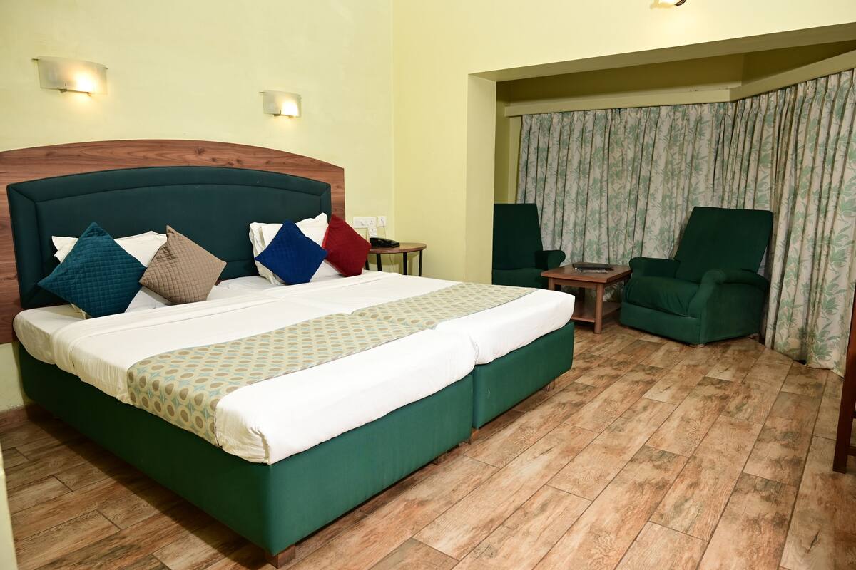 A hotel room with a large bed, a green armchair, a small table, and a curtain.