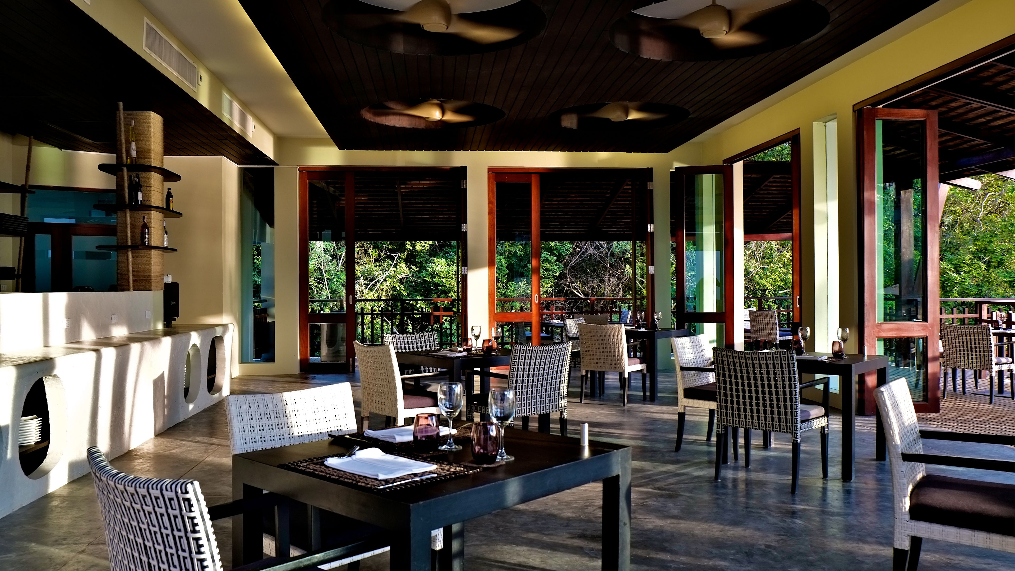 An outdoor dining area with tables and chairs, large windows, and a view of greenery.