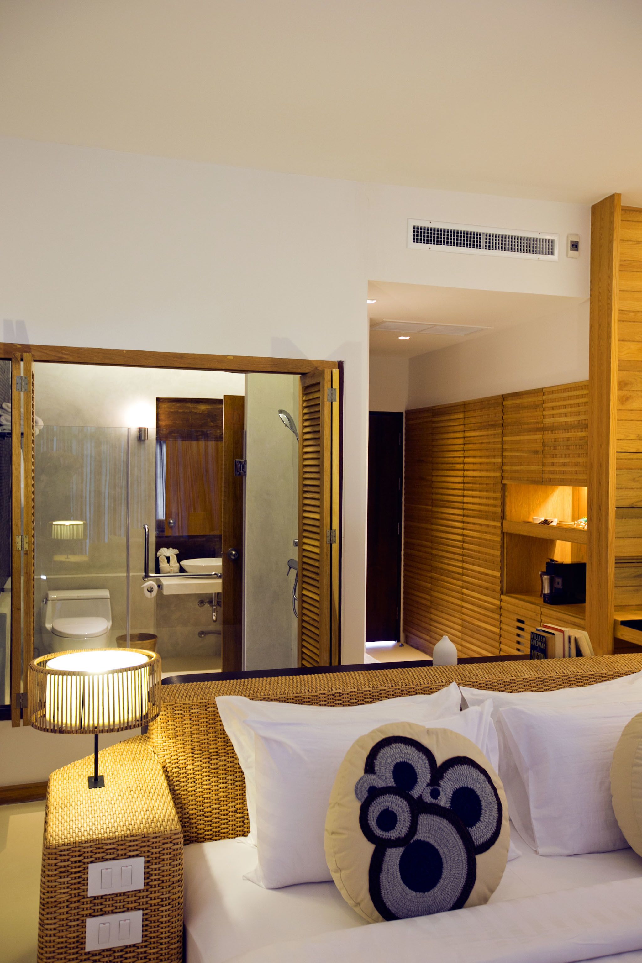 A hotel room with a bed, a bedside lamp, a wicker side table, a mirror, and a wooden wardrobe.
