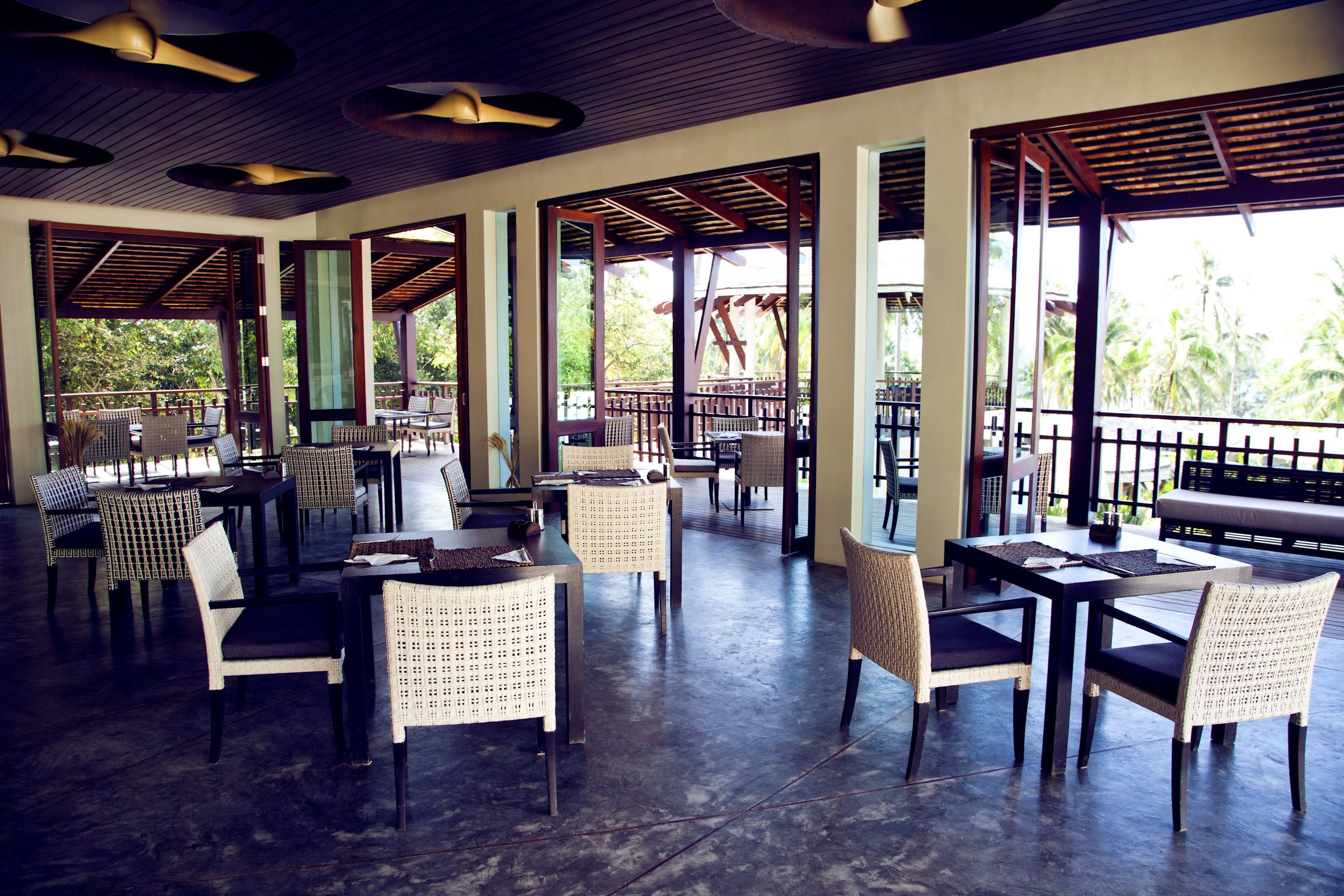 Ein Restaurant im Freien mit hohen Decken, großen Fenstern und unterschiedlichen Sitzmöglichkeiten, darunter Tische und Stühle.