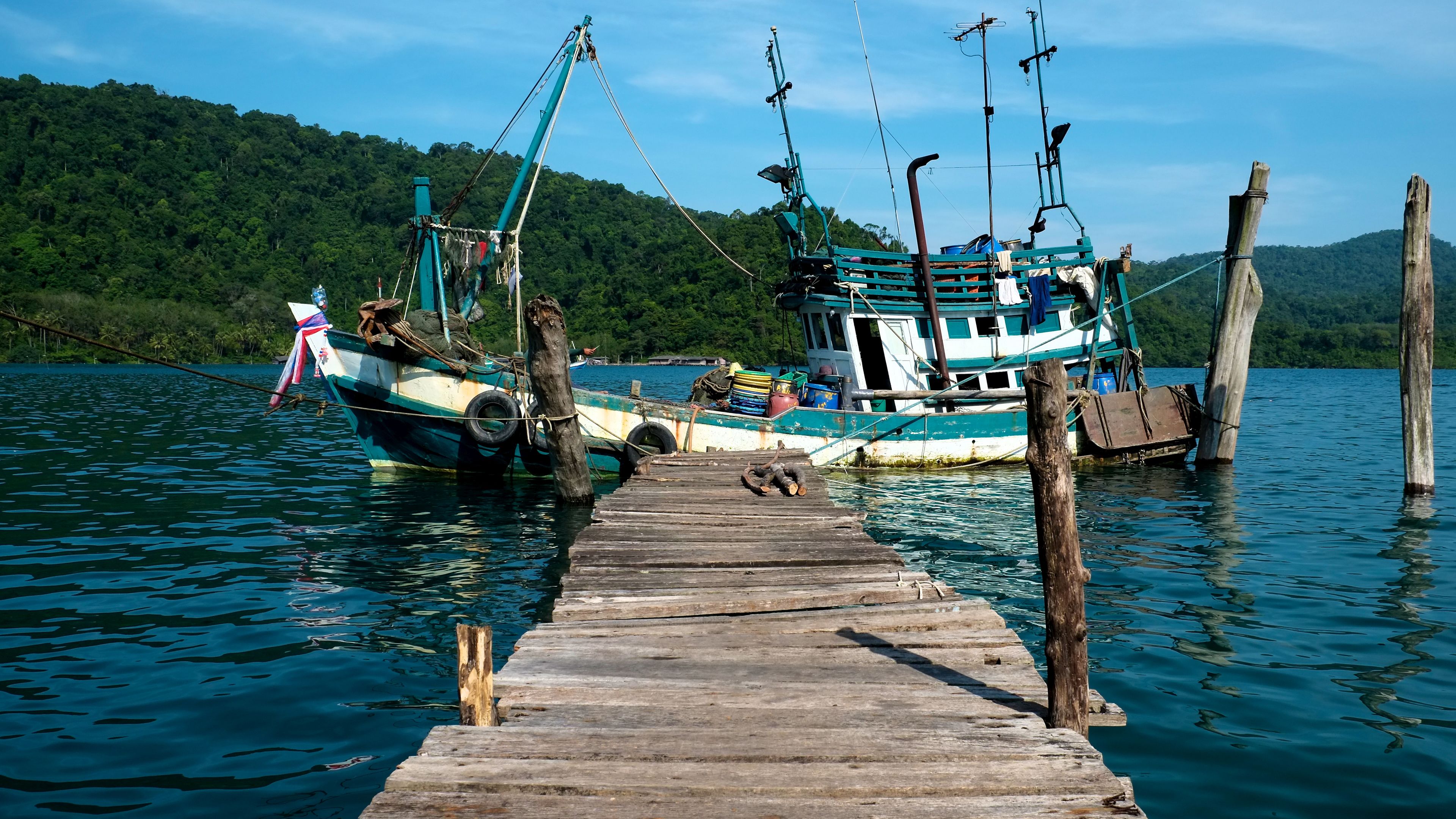 A wooden pier leading to a boat docked at a calm lake with a forested hill in the background.