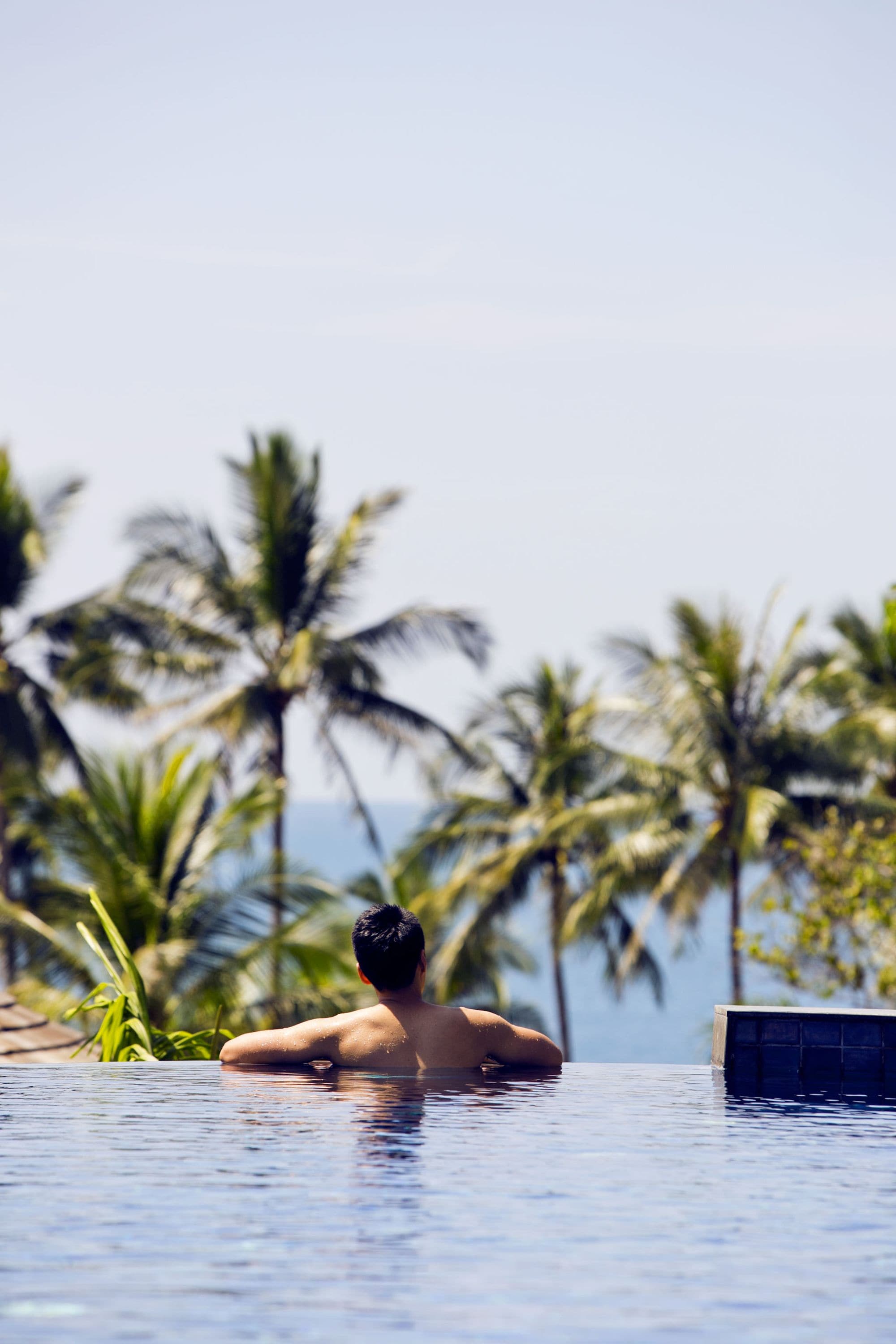 Un hombre en una piscina con vista a palmeras y al océano.