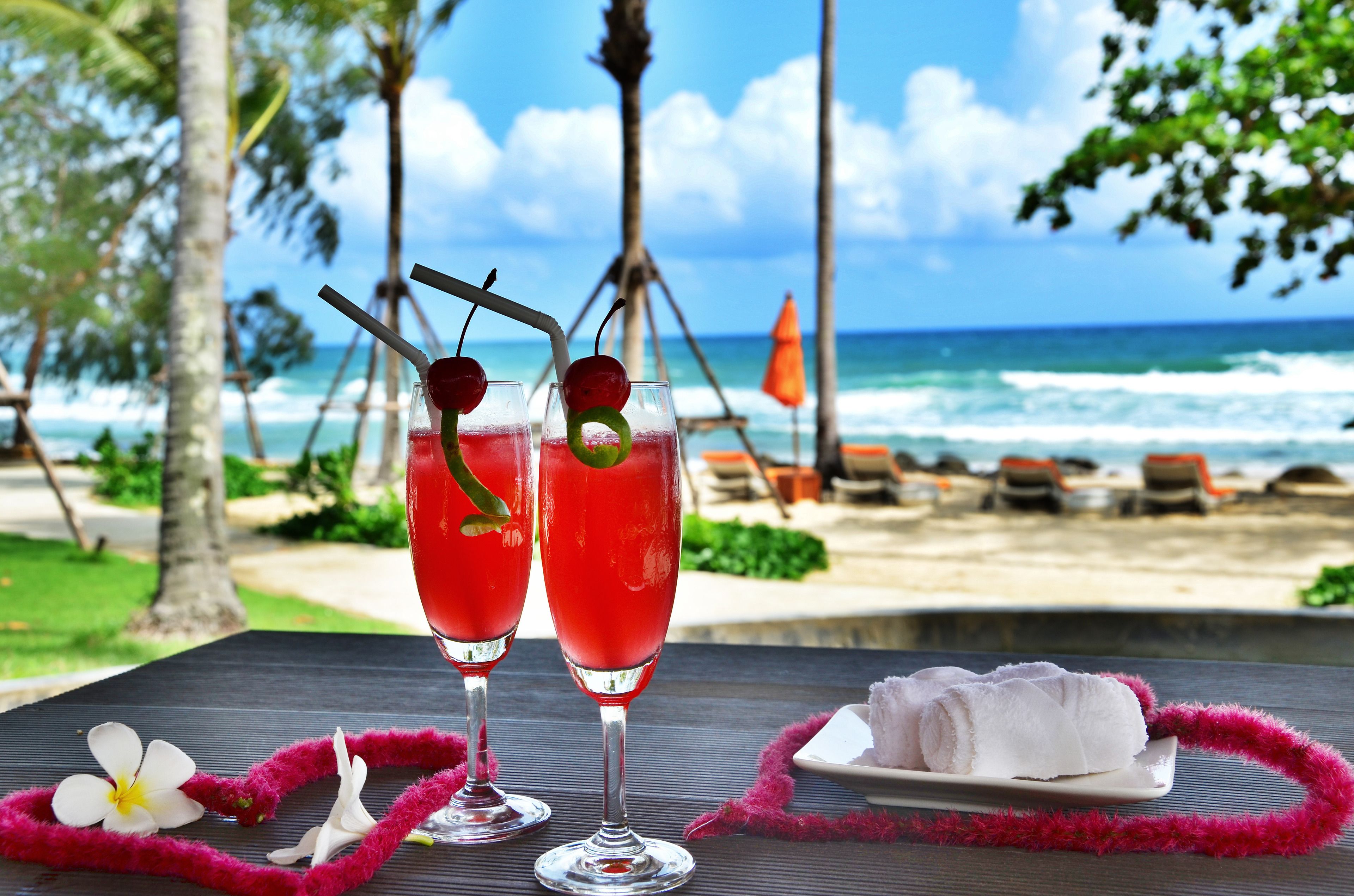 Two cocktails on a table with a beach view, a plate of towels, and a lei.